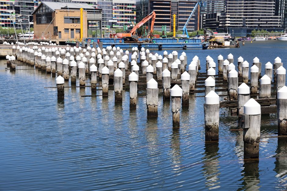 A row of wooden mooring posts extending into a body of water, each topped with white, conical protective caps. The posts are weathered, showing signs of age with darkened, rough textures. In the background, a small floating platform or barge is visible, equipped with crane and construction machinery, suggesting some form of dockside activity. Surrounding the platform are modern high-rise buildings with glass facades, indicating an urban waterfront setting. The water is calm, reflecting the posts and buildings, with gentle ripples visible on the surface. The environment appears to be a mixing of a working waterfront or marina area, with infrastructure suitable for boat mooring or small-scale industrial work. The scene is lit by natural daylight, providing clear visibility of the textures and colors, which include the neutral tones of the wooden posts and the reflective surfaces of the water and glass buildings. The setting underscores a functional, on-site maritime environment that may be associated with local port or dockyard activities, indirectly linking to practices of onsite disposal or waste handling relative to urban maritime operations associated with waste clearance services like those offered by Waste Clearance Docklands.