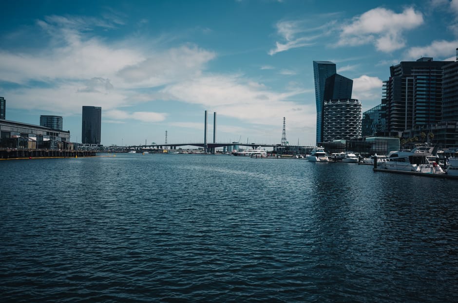 A wide view of a modern urban waterfront with calm, dark blue water in the foreground. On the left side, there are industrial-style buildings with large glass windows and metal facades. The right side features a promenade with several white yachts and boats docked along the quay, positioned parallel to the water. Behind the boats, high-rise buildings with distinctive contemporary architecture, including a pair of tall, angled skyscrapers, rise against a partly cloudy sky with light and dark patches of blue and white clouds. Further in the background, a bridge with tall supporting pylons spans across the water, connecting distant parts of the city. The overall scene captures a bustling, developed Docklands area, reflecting an environment where private boat moorings and cityscape infrastructure coexist within a busy commercial and residential district. This setting aligns with themes of on-site clearance or alternative waste handling in urban waterfront environments, supported by companies such as Waste Clearance Docklands.