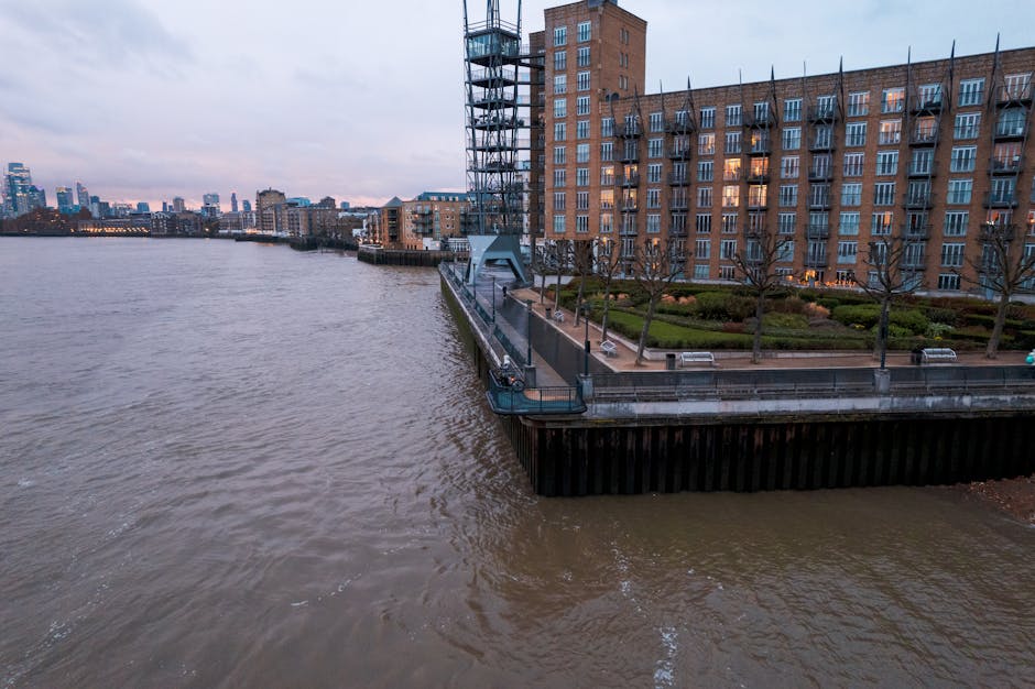 A view of a riverfront promenade along a residential building in Docklands, with a brick multi-storey apartment complex visible on the right side, featuring numerous windows with some illuminated. The promenade is paved and includes small, leafless trees evenly spaced along the edge, with a neatly maintained garden bed and benches. A metal railing runs along the waterfront, and a small circular dock or platform extends over the water, supported by dark vertical fencing at its base. In the background, there are various high-rise buildings and office towers in a distant city skyline, under a cloudy, overcast sky, suggesting late afternoon or early evening. The river water appears brown and slightly choppy. The image captures the urban environment typical of private waterfront properties, with a subtle visual connection to services like rubbish removal or on-site clearance carried out by firms such as Waste Clearance Docklands, focusing on maintaining tidy and safe residential areas.
