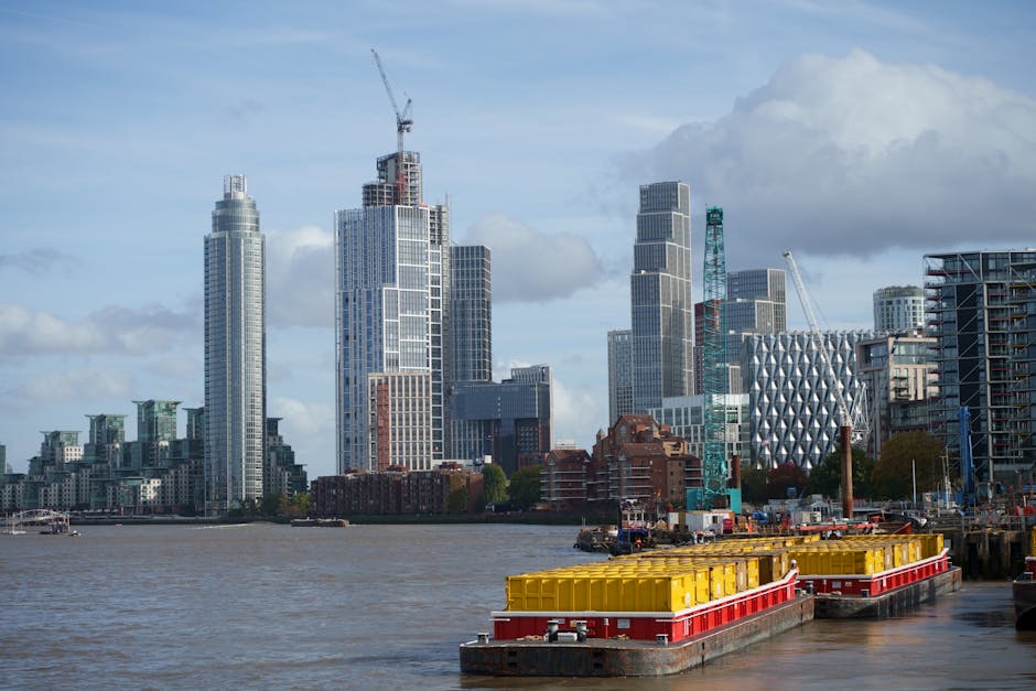 A view of a riverfront promenade along a residential building in Docklands, with a brick multi-storey apartment complex visible on the right side, featuring numerous windows with some illuminated. The promenade is paved and includes small, leafless trees evenly spaced along the edge, with a neatly maintained garden bed and benches. A metal railing runs along the waterfront, and a small circular dock or platform extends over the water, supported by dark vertical fencing at its base. In the background, there are various high-rise buildings and office towers in a distant city skyline, under a cloudy, overcast sky, suggesting late afternoon or early evening. The river water appears brown and slightly choppy. The image captures the urban environment typical of private waterfront properties, with a subtle visual connection to services like rubbish removal or on-site clearance carried out by firms such as Waste Clearance Docklands, focusing on maintaining tidy and safe residential areas.