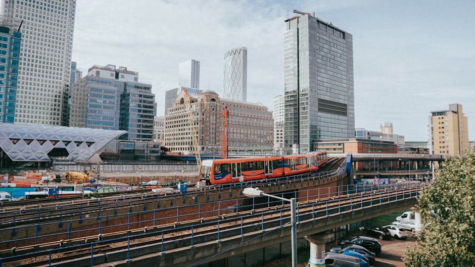 The image depicts an urban high street scene with a modern train crossing an elevated railway track in the foreground. The train is orange with black and white accents, positioned centrally on the tracks, suggesting on-site or private transportation. Behind the railway, a variety of tall, contemporary office buildings and skyscrapers with glass facades reflect diffused daylight in a mix of blue, grey, and beige tones. The buildings vary in architectural style, with some featuring grid-like window patterns and others with curved or angular designs. To the right, a parking lot filled with parked cars is partially visible beneath the railway, with some greenery and trees bordering the lot. The sky appears overcast with a light, even cloud cover, providing soft illumination across the scene. The overall setting suggests a busy, developed city area, with potential links to local waste and rubbish management services like Waste Clearance Docklands, capturing a typical scene of infrastructure and urban activity that may relate to the logistics of rubbish removal or transportation.