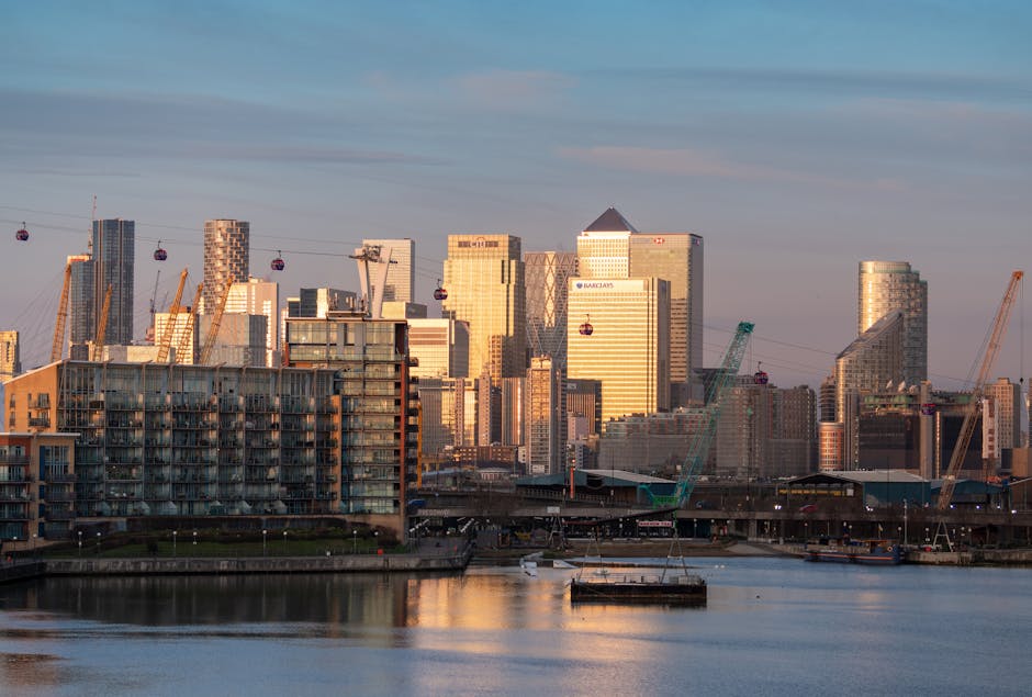 A panoramic view of a modern city skyline during sunset, featuring high-rise office buildings with reflective glass facades, some with distinct geometric designs and varying heights, set against a partly cloudy sky. Several construction cranes are visible among the clusters of tall structures, indicating ongoing development, with cable cars crossing in the foreground above the river. At the riverbank, there are mid-rise residential buildings with balconies and textured exteriors, situated near a paved promenade. The water in the river is calm, reflecting the warm hues of the setting sun and the silhouettes of the buildings, creating a tranquil atmosphere. The scene suggests a vibrant urban area with active construction activity, and such a setting could relate to areas where private waste disposal and on-site clearance services may operate, supported by companies like Waste Clearance Docklands, to handle rubbish associated with ongoing developments or urban upgrades.
