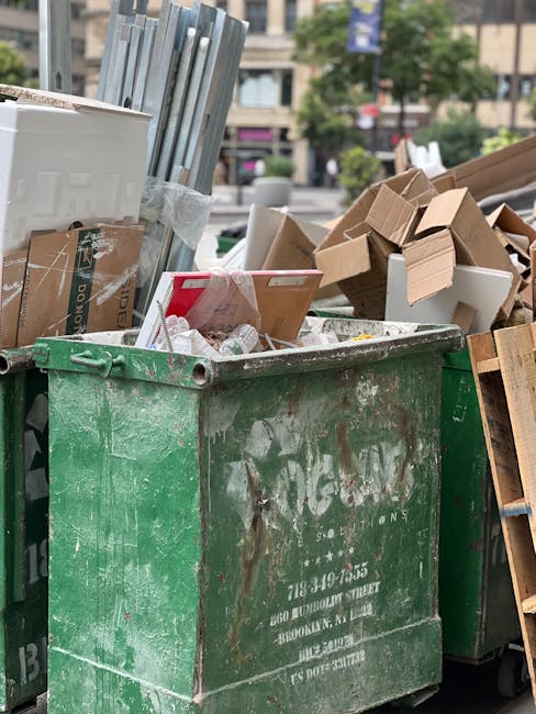 A large, weathered green waste skip filled with various types of discarded materials, including flattened cardboard boxes, some with printed text and logos, white foam packaging pieces, and smaller cardboard fragments. The skip is situated outdoors on a paved area, with additional cardboard boxes stacked nearby, some leaning against the skip's sides. In the background, blurred trees and multi-story buildings can be seen, indicating an urban environment. The scene suggests an on-site clearance or waste disposal process, demonstrating private waste handling typical of services like those provided by Waste Clearance Docklands in the East London area. The lighting is natural, with slight shadows cast over the waste, emphasizing the textures of the cardboard, foam, and the metal surface of the skip. This visual context aligns with an alternative approach to rubbish collection, focusing on independent waste removal solutions suited to local businesses or residents managing their own rubbish disposal outside of standard council services.