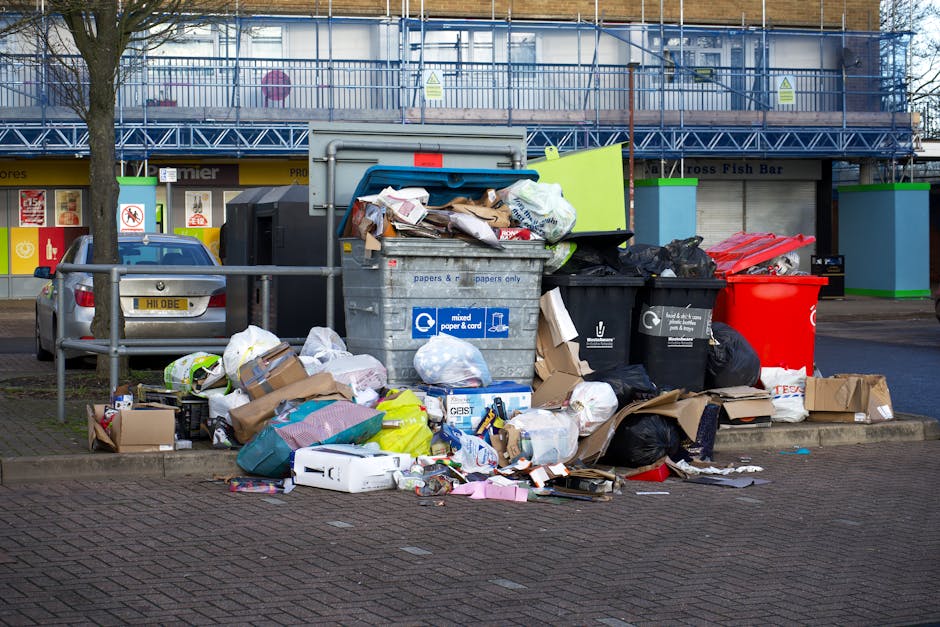 A collection of overflowing rubbish bins and scattered waste on a paved urban sidewalk area near a metal railing, with a grey metal recycling container filled with mixed paper and cardboard on the front. The waste includes black bin bags, flattened cardboard boxes, paper wrappings, plastic packaging, and other household rubbish. The scene is set against a commercial building with large windows and blue scaffolding, and a storefront with signage visible in the background. A parked car with a partially visible license plate is situated close to the waste area, indicating proximity to a parking lot. The rubbish appears to be accumulated for disposal or for private collection, reflecting a typical location where waste clearance services such as those provided by Waste Clearance Docklands might be involved in removing accumulated refuse from an area that has not been cleared recently.
