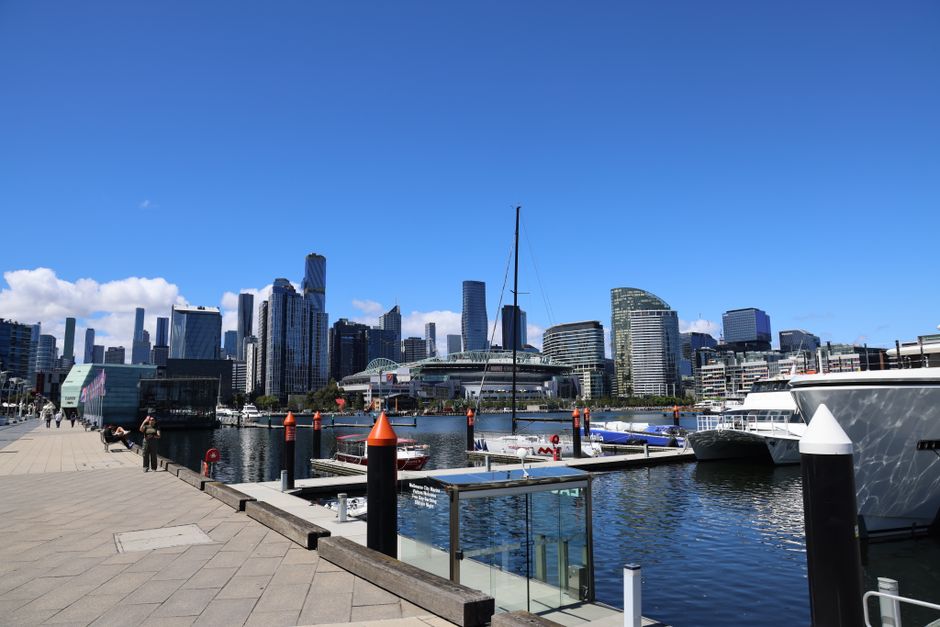 A city waterfront scene featuring a clear blue sky with a few scattered white clouds. In the foreground, a paved promenade runs parallel to the water, with a few pedestrians walking or standing near the edge. Several boats and yachts are docked along the marina, including a large white yacht on the right side and a smaller vessel with a dark hull and white superstructure. The marina is protected by black and orange navigation posts along the quay. Beyond the water, the skyline is dominated by modern high-rise buildings of varying heights and architectural styles, primarily composed of glass facades reflecting sunlight, with some featuring curved or angular designs. The area conveys a vibrant urban environment, typical of a prominent docklands district, with the scene suggesting an active maritime setting suitable for private and commercial vessel mooring, complementing alternative waste disposal methods for nearby buildings and facilities managed by Waste Clearance Docklands and other services specializing in rubbish and waste removal in the area.